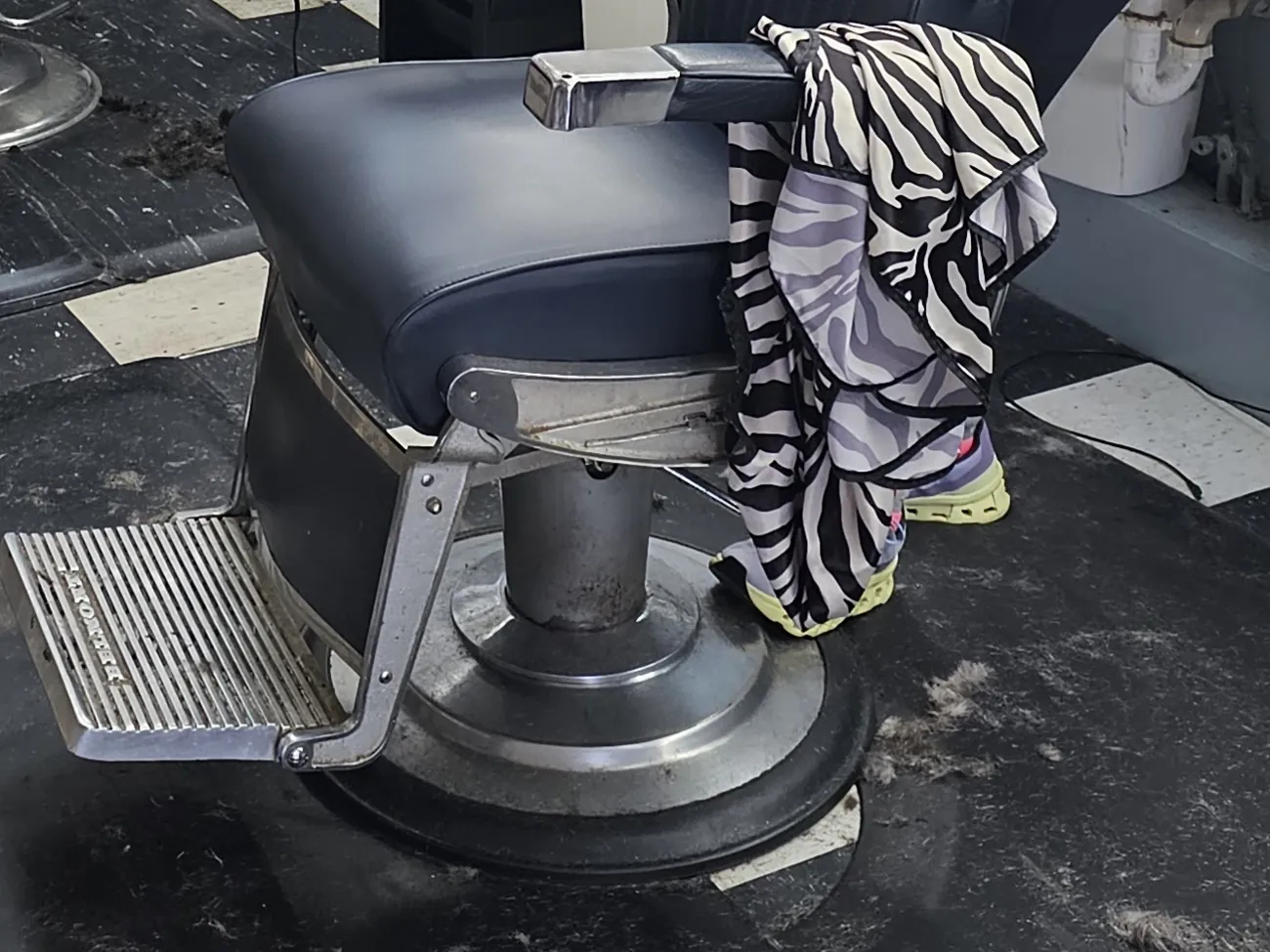 A vintage barber chair with a navy seat and chrome footrest on a black-and-white checkerboard floor, a zebra-print cape draped over the arm