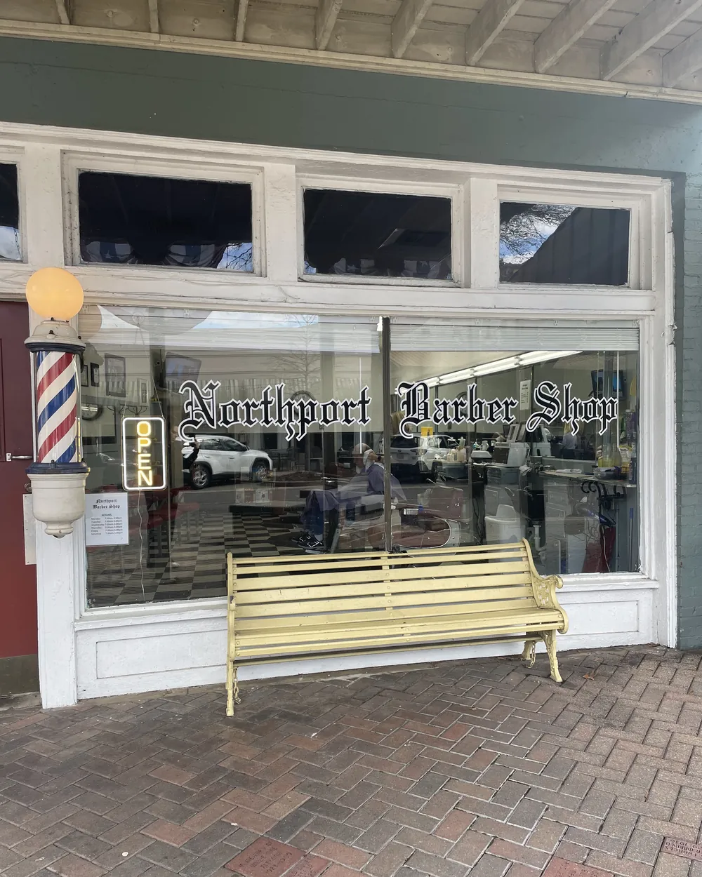 The storefront on Main Avenue: a striped barber pole and neon OPEN sign beside a big window lettered Northport Barber Shop, with a yellow bench out front on a brick sidewalk
