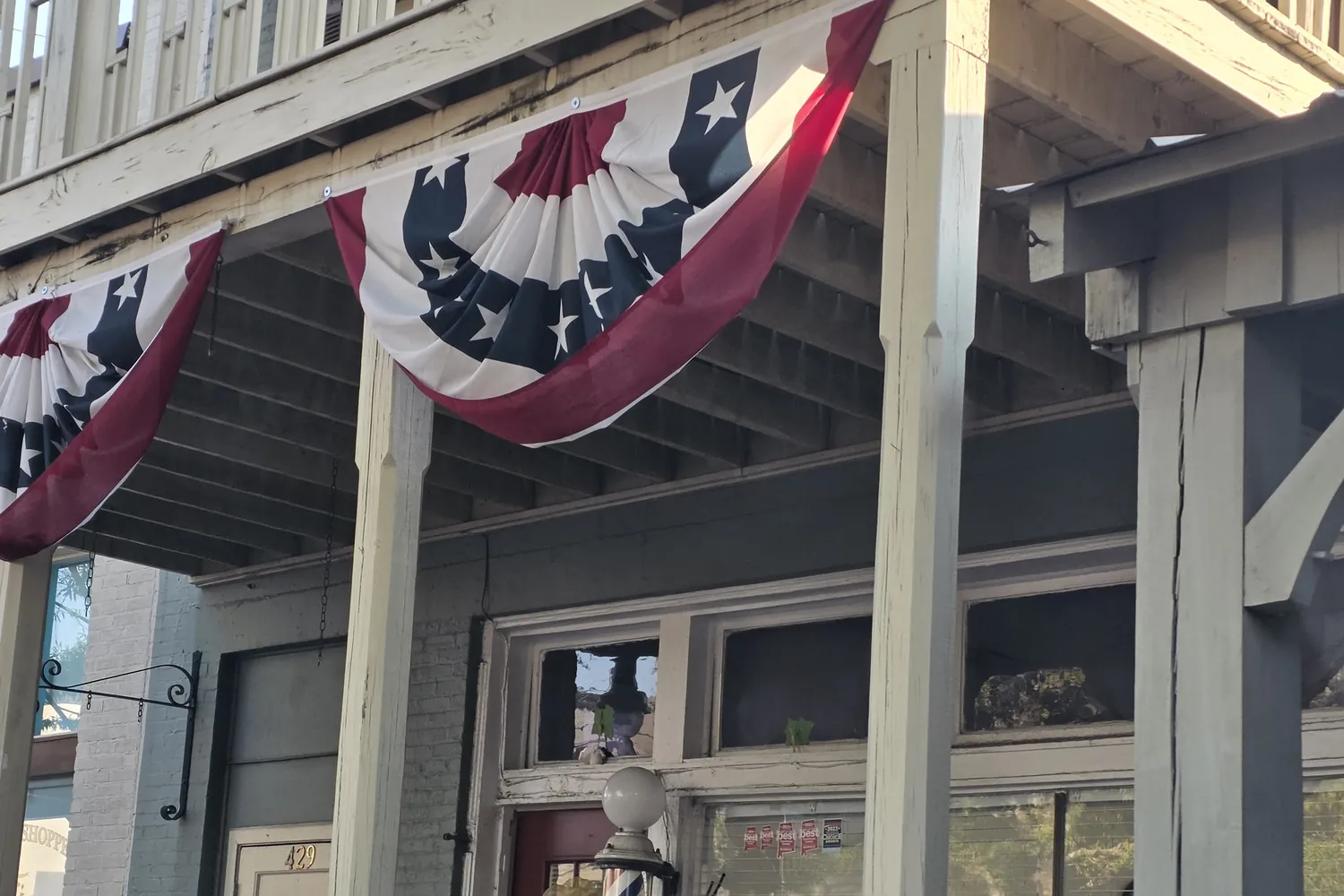 The shop front under the upstairs porch, with red, white and blue flag bunting hung from the railing and a barber pole by the door