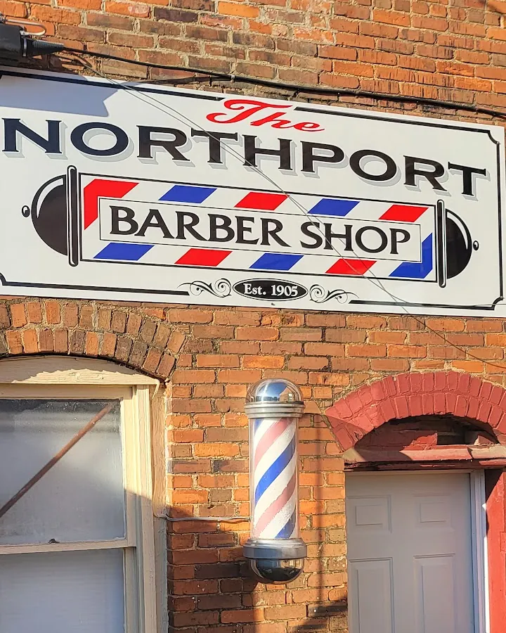 Hanging sign on a red brick wall reading The Northport Barber Shop, Est. 1905, with a small striped barber pole and a white side door below it