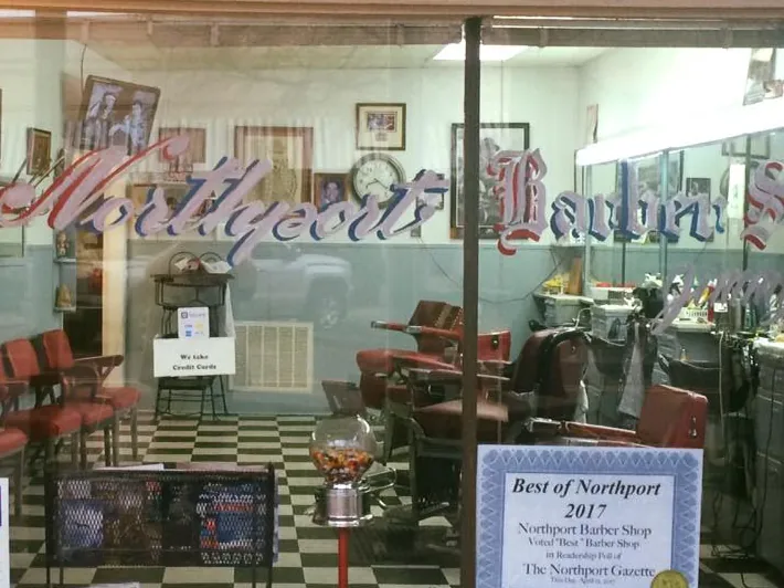 Through the shop window: red waiting chairs, framed photos on the wall, a gumball machine and a Best of Northport 2017 certificate from The Northport Gazette